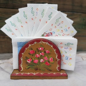 May include: Wooden napkin holder with a floral design painted on the front. The holder is painted in shades of brown, red, and green. The holder is holding a stack of white napkins with musical notes printed on them.