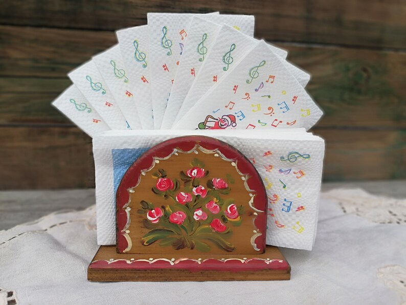 May include: Wooden napkin holder with a floral design painted on the front. The holder is painted in shades of brown, red, and green. The holder is holding a stack of white napkins with musical notes printed on them.