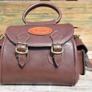 May include: A brown leather Wrangler handbag with a top handle and a detachable shoulder strap. The bag features two front buckles and a small leather patch with the Wrangler logo.