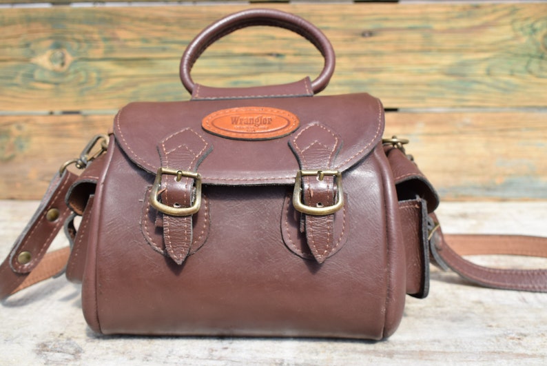 May include: A brown leather Wrangler handbag with a top handle and a detachable shoulder strap. The bag features two front buckles and a small leather patch with the Wrangler logo.