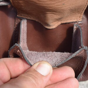 May include: Close-up of a brown leather pouch with a flap. The pouch is open, revealing a lighter brown interior. The leather appears to be of high quality, with visible stitching and a textured surface.
