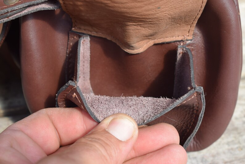 May include: Close-up of a brown leather pouch with a flap. The pouch is open, revealing a lighter brown interior. The leather appears to be of high quality, with visible stitching and a textured surface.
