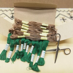 May include: A box of green embroidery floss with brown spools of thread inside. The box is open and the floss is spilling out. A pair of silver scissors is lying on top of the box.