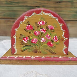 May include: A wooden napkin holder with a hand-painted design of pink roses and green leaves on a yellow background. The holder has a red trim with white accents.