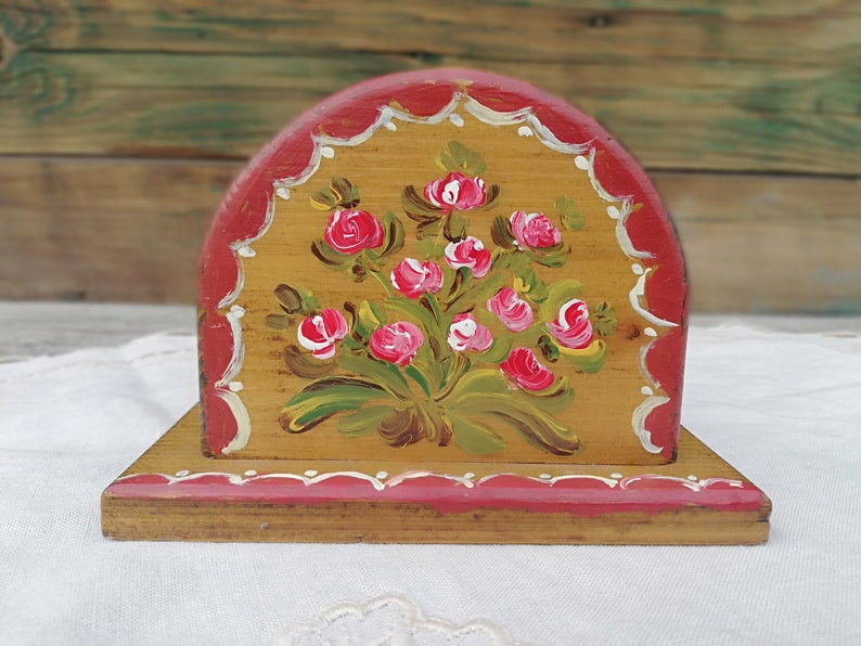 May include: A wooden napkin holder with a hand-painted design of pink roses and green leaves on a yellow background. The holder has a red trim with white accents.