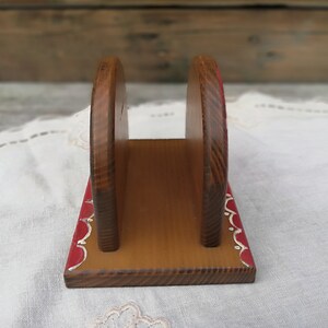 May include: Wooden napkin holder with a red and white painted design. The holder has a rectangular base and two vertical sides that form a slot for napkins.