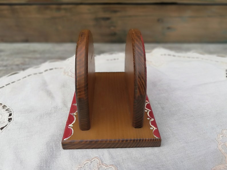 May include: Wooden napkin holder with a red and white painted design. The holder has a rectangular base and two vertical sides that form a slot for napkins.