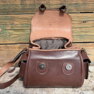 May include: Open brown leather satchel bag with a flap closure. The bag has a shoulder strap and two small pockets on the sides. The interior is lined with a darker fabric. The bag is sitting on a wooden surface.