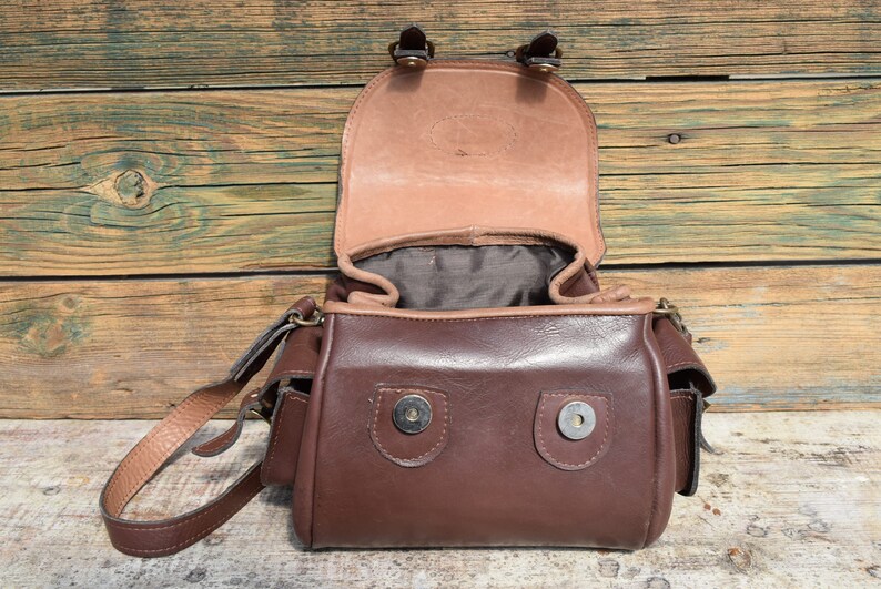 May include: Open brown leather satchel bag with a flap closure. The bag has a shoulder strap and two small pockets on the sides. The interior is lined with a darker fabric. The bag is sitting on a wooden surface.