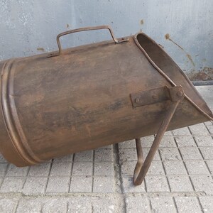 May include: A rusty metal coal scuttle with a handle and a single leg. The scuttle is sitting on a tiled floor.