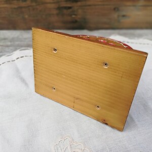 May include: The bottom of a wooden napkin holder with a light brown finish. The holder has four small screws on the bottom.