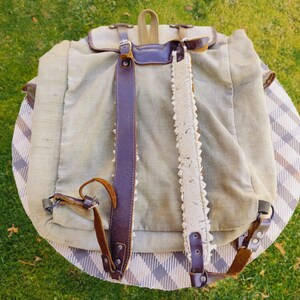 Op de afbeelding: Een vintage canvas rugzak met bruine leren banden en een witte kantversiering. De rugzak heeft een klep sluiting en een gesp.