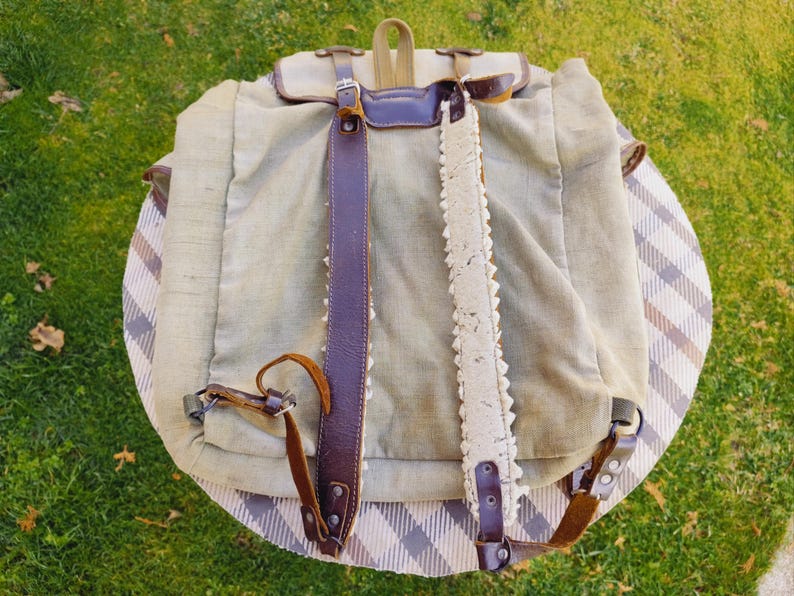 Op de afbeelding: Een vintage canvas rugzak met bruine leren banden en een witte kantversiering. De rugzak heeft een klep sluiting en een gesp.