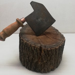 May include: A rusty, metal cleaver with a wooden handle sits on a wooden stump. The cleaver has a copper accent on the handle.