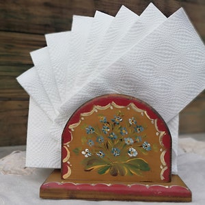 May include: A wooden napkin holder with a hand-painted floral design in shades of blue, green, and yellow. The holder has a red base and a curved top.
