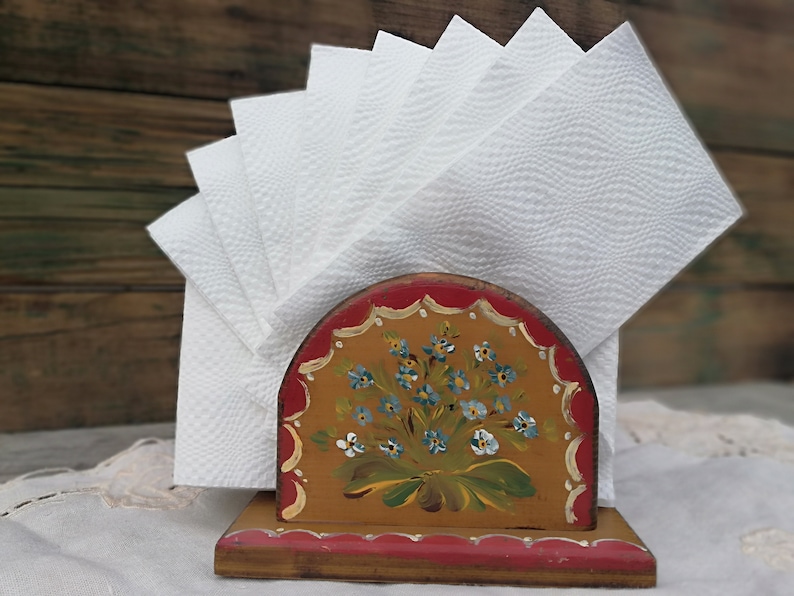 May include: A wooden napkin holder with a hand-painted floral design in shades of blue, green, and yellow. The holder has a red base and a curved top.