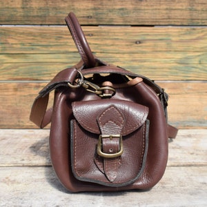 May include: A brown leather satchel with a front pocket secured by a brass buckle. The bag has a top handle and a shoulder strap. The leather appears to be textured, and the bag is set against a wooden background.