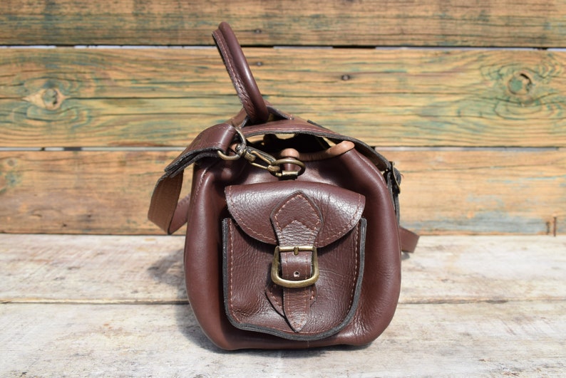 May include: A brown leather satchel with a front pocket secured by a brass buckle. The bag has a top handle and a shoulder strap. The leather appears to be textured, and the bag is set against a wooden background.