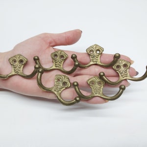 May include: Five gold-toned metal double hooks with ornate designs. The hooks are arranged in a semi-circle.