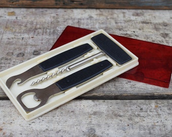 May include: A white plastic case with a red plastic lid containing a set of three black handled tools: a bottle opener, a corkscrew, and a bottle stopper.