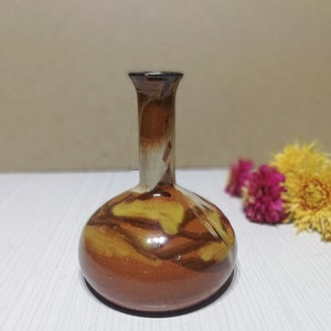 Puede incluir: Un pequeño jarrón de cerámica marrón con una base ancha y un cuello estrecho. El jarrón tiene un esmalte moteado marrón, amarillo y blanco.