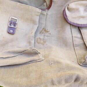 Op de afbeelding: Een beige canvas tas met bruine leren afwerking en een zilveren gesp. De tas heeft een paar bruine vlekken.