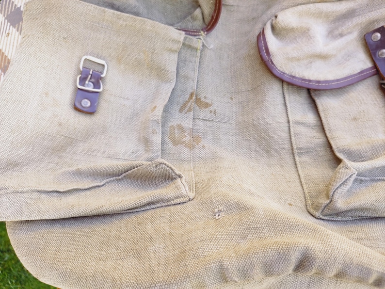 Op de afbeelding: Een beige canvas tas met bruine leren afwerking en een zilveren gesp. De tas heeft een paar bruine vlekken.