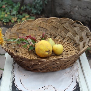 Puede incluir: Una cesta de mimbre tejida con un esquema de color marrón y beige, llena de un membrillo amarillo, una pera verde y una manzana amarilla. La cesta está sentada sobre un encaje blanco sobre una superficie blanca.