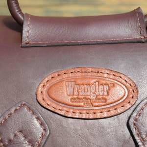 May include: Close-up of a brown leather bag with a handle. The bag features a stitched leather patch with the "Wrangler" logo. The bag has a dark brown color and a textured surface.