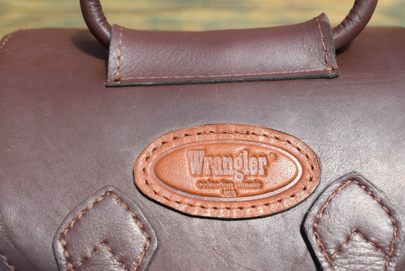 May include: Close-up of a brown leather bag with a handle. The bag features a stitched leather patch with the "Wrangler" logo. The bag has a dark brown color and a textured surface.