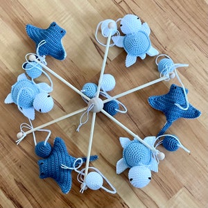 May include: A wooden baby mobile with blue and white crocheted sea creatures, including a starfish, a turtle, and a stingray. The mobile is suspended from a wooden ring and has white and blue balls hanging from it.