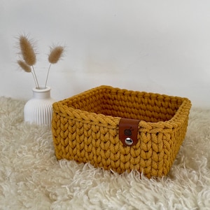Op de afbeelding: Een mosterdgele gehaakte opbergmand met een bruin leren handvat en een klein zwart logo. De mand staat op een wit, pluche oppervlak.