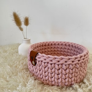 Op de afbeelding: Een roze gehaakte mand met een bruin leren handvat. De mand staat op een wit, pluche oppervlak.