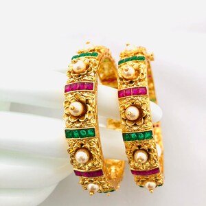 May include: Two gold bangles with intricate floral designs, adorned with green and pink gemstones and white pearls.