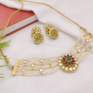 May include: A gold choker necklace with a large, round pendant featuring a red and green gemstone design. The necklace is made of multiple strands of white beads and has a gold clasp. The necklace comes with a pair of matching earrings with a similar design.