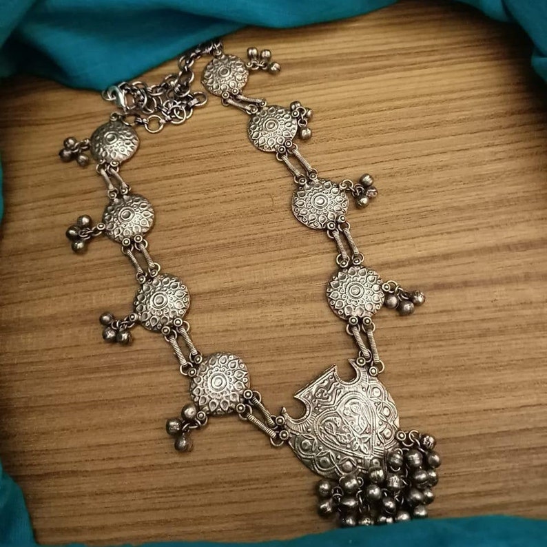 May include: A silver necklace with a large, ornate pendant and multiple smaller circular pendants. The necklace is made of metal and has a textured finish.