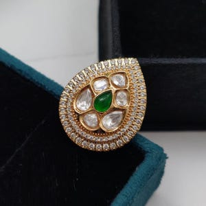 May include: A gold teardrop-shaped pendant with a green gemstone surrounded by smaller white gemstones. The pendant is set in a gold frame with a row of smaller white gemstones around the edge.