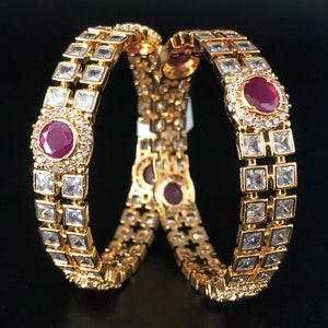 May include: Two gold bangles with a row of clear square stones and a larger red stone in the center of each bangle.