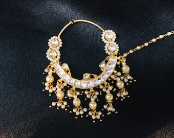 May include: A gold nose ring with a pearl and gemstone design. The ring has a chain attached to it, which is also adorned with pearls.