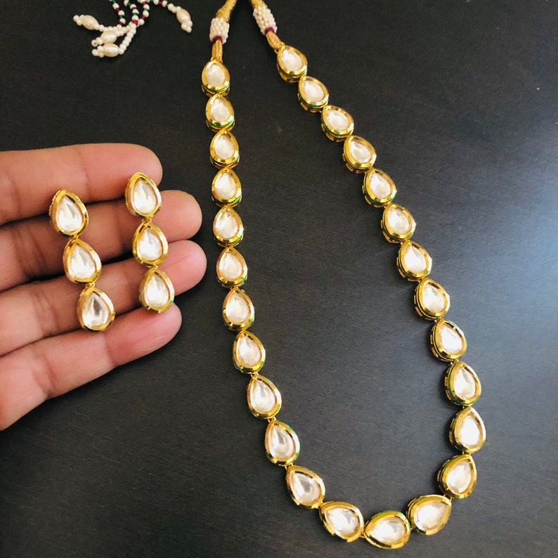 May include: A gold necklace and earrings set with teardrop-shaped white stones. The necklace is made of a gold chain with a series of teardrop-shaped stones.