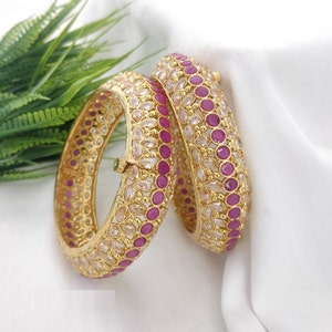 May include: Two gold bangles with intricate designs. One bangle is decorated with pink and white stones, while the other is decorated with white stones.