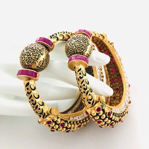 May include: Two gold bangles with intricate black enamel designs and pink gemstones. The bangles are decorated with small round beads and have a decorative ball at the end.