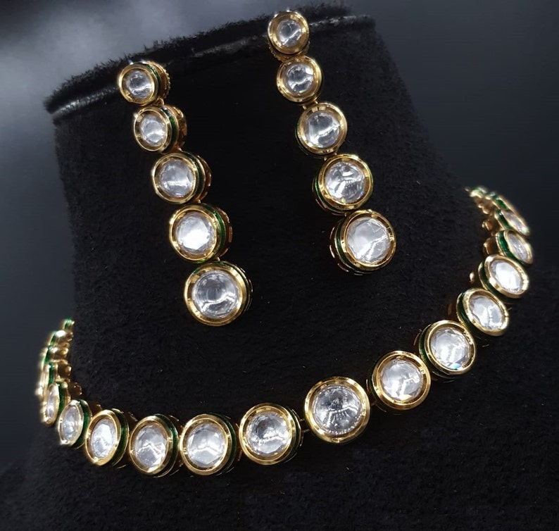 May include: A gold necklace and earrings set with a row of clear round stones with green accents.