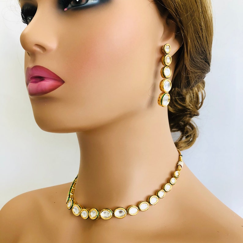 May include: A gold necklace and earrings set with oval-shaped clear stones surrounded by green accents. The necklace is a choker style and the earrings are long and dangle.