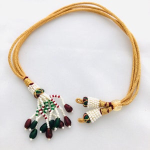 Puede incluir: Un collar de oro con un borla hecha de cuentas blancas, verdes y rojas. La borla tiene pequeñas cuentas de color rojo oscuro y verde en el extremo. El collar está hecho de un cordón trenzado.