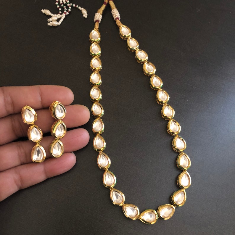 May include: A gold necklace and earrings set featuring teardrop-shaped clear stones. The necklace is made of a chain with a series of teardrop-shaped stones. The earrings are a pair of teardrop-shaped stones.