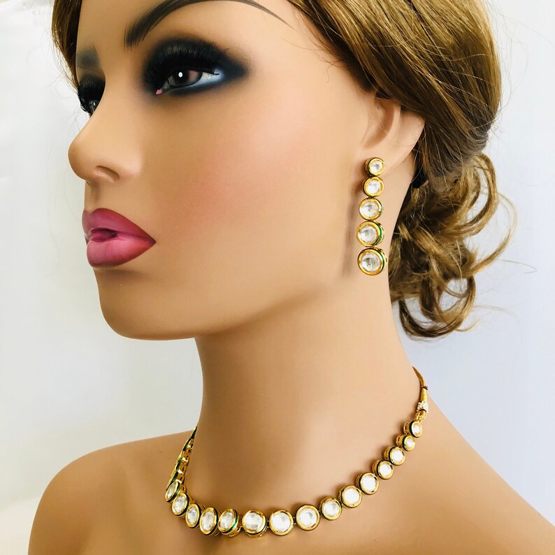 May include: A gold necklace and earrings set with round white stones and green accents. The necklace is a choker style with a row of stones.