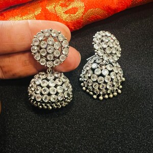 May include: A pair of silver earrings with a round, multi-faceted design. The earrings are adorned with clear stones and have a dangling, bell-shaped bottom.