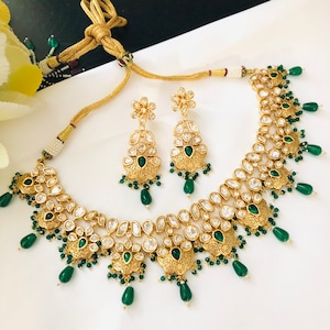May include: A gold necklace and earrings set with green and white stones. The necklace has a layered design with multiple tiers of stones. The earrings are dangle style with a similar design to the necklace.