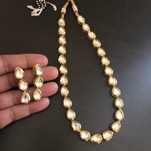 May include: A gold necklace with a teardrop-shaped pendant and matching earrings. The necklace is made of gold and has a white stone in the centre of each pendant. The earrings are also made of gold and have a white stone in the centre of each pendant.